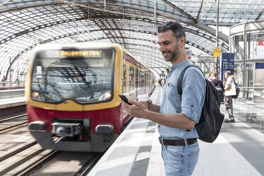 Lächelnder Geschäftsmann am Bahnhof, der auf den Zug wartet und auf sein Smartphone schaut, Berlin, Deutschland - WPEF02098