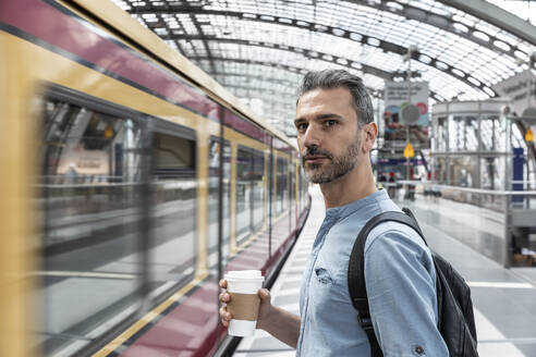 Mann mit Kaffee zum Mitnehmen am Bahnhof, während ein Zug einfährt, Berlin, Deutschland - WPEF02099