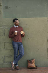 Junger Mann mit einem Cheeseburger und einem Kaffee zum Mitnehmen, an die Wand gelehnt - VPIF01629