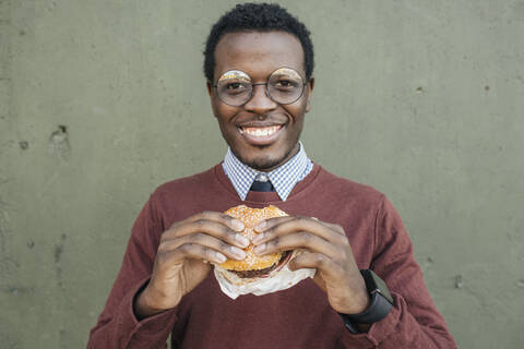 Junger Mann isst Cheeseburger, lächelnd, lizenzfreies Stockfoto