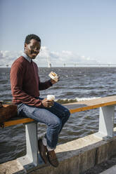 Junger Mann sitzt auf der Reling am Meer, isst Hamburger, trinkt Kaffee - VPIF01641