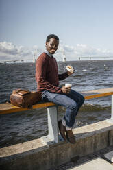 Junger Mann sitzt auf der Reling am Meer, isst Hamburger, trinkt Kaffee - VPIF01642