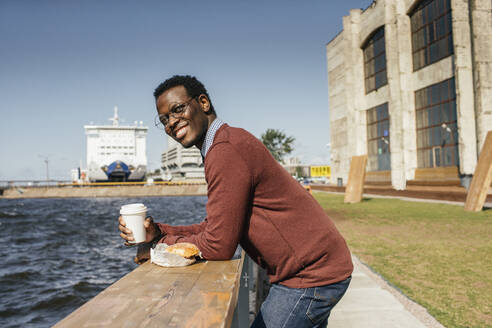 Young man looking at the sea, with eating hamburgerand coffee on railing - VPIF01647