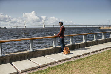 Young man sitting on railing at the sea, eating hamburger, drinking coffee - VPIF01648