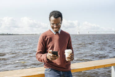 Young man standing at the sea, holding coffee, using smartphone - VPIF01653