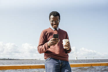 Young man standing at the sea, holding coffee, using smartphone - VPIF01654