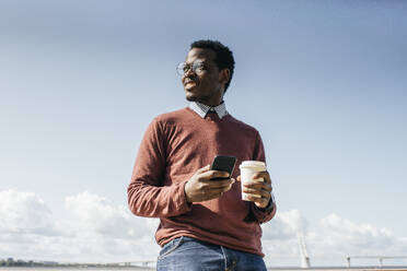 Young man standing at the sea, holding coffee, using smartphone - VPIF01655