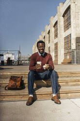 Junger Mann sitzt auf einer Treppe in der Stadt, isst einen Hamburger und trinkt Kaffee - VPIF01663