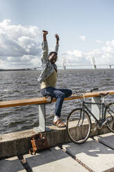 Junger Mann mit Fahrrad, sitzt auf einer Reling am Meer und streckt sich - VPIF01709