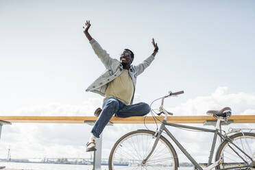 Junger Mann mit Fahrrad, der auf einer Reling am Meer sitzt und so tut, als würde er fliegen - VPIF01711