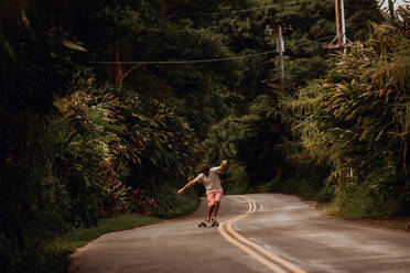 Mid adult male skateboarder skateboarding along rural road with arms outstretched, Haiku, Hawaii, USA - ISF22597