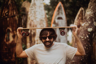 Mittlerer Erwachsener, männlicher Skateboarder, der ein Skateboard auf dem Kopf trägt, neben einer alten Surfbrettstruktur im Park, Porträt, Haiku, Hawaii, USA - ISF22700