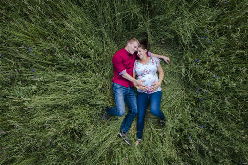 Pregnant woman and man holding baby belly, lying on meadow - HMEF00661