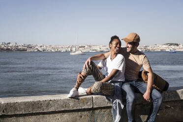 Glückliches junges Paar sitzt auf einer Mauer am Wasser, Lissabon, Portugal - UUF19062