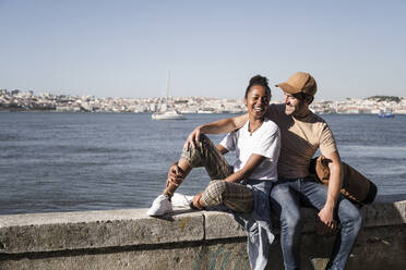 Glückliches junges Paar sitzt auf einer Mauer am Wasser, Lissabon, Portugal - UUF19063