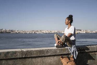 Junge Frau sitzt auf einer Mauer am Wasser, Lissabon, Portugal - UUF19067