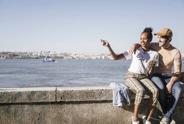 Glückliches junges Paar sitzt auf einer Mauer am Wasser, Lissabon, Portugal - UUF19072