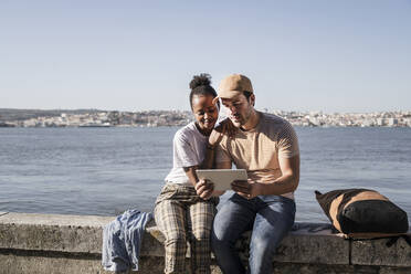 Junges Paar sitzt auf einer Mauer am Wasser und benutzt ein Tablet, Lissabon, Portugal - UUF19073