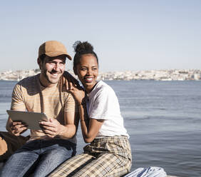 Happy young couple sitting at the waterfront with a tablet, Lisbon, Portugal - UUF19075