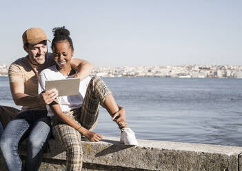 Glückliches junges Paar sitzt auf einer Mauer am Wasser und benutzt ein Tablet, Lissabon, Portugal - UUF19076
