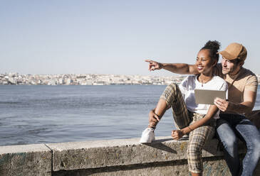 Young couple sitting on a wall at the waterfront with a tablet, Lisbon, Portugal - UUF19077