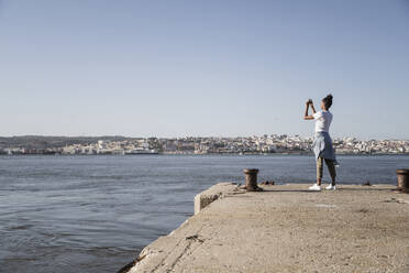 Junge Frau steht am Pier am Wasser und macht ein Handyfoto, Lissabon, Portugal - UUF19091