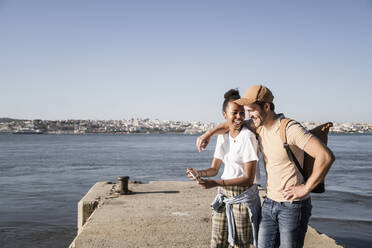 Happy young couple using cell phone on pier at the waterfront, Lisbon, Portugal - UUF19104