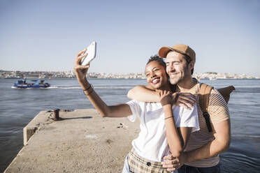 Happy young couple taking a selfie on pier at the waterfront, Lisbon, Portugal - UUF19106