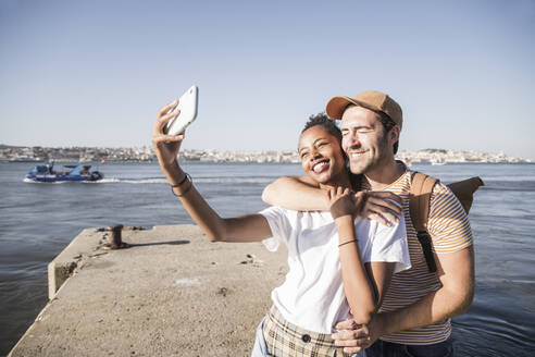 Happy young couple taking a selfie on pier at the waterfront, Lisbon, Portugal - UUF19106