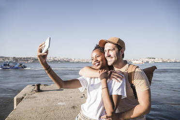 Happy young couple taking a selfie on pier at the waterfront, Lisbon, Portugal - UUF19107