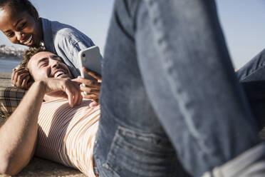 Glückliches junges Paar liegt auf dem Pier und benutzt ein Mobiltelefon - UUF19110