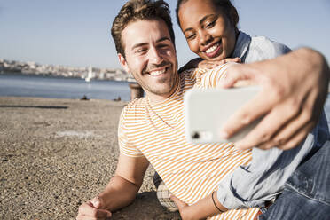 Happy young couple taking a selfie on pier at the waterfront, Lisbon, Portugal - UUF19116