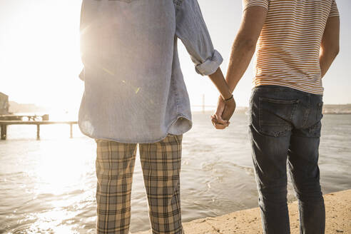 Close-up of couple holding hands at the waterfront at sunset - UUF19125