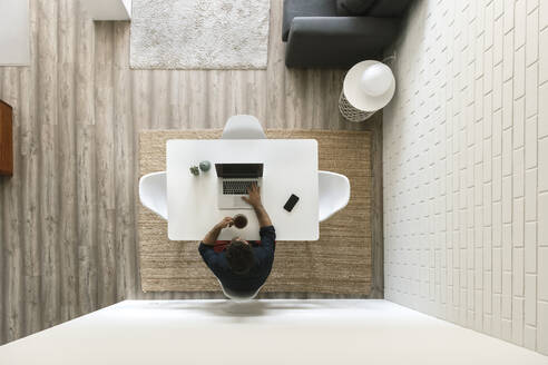 Man sitting at table in modern loft using laptop while drinking coffee, top view - AHSF01029