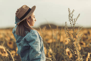 Junge Frau mit Hut und Jeansjacke in einem Sonnenblumenfeld im Abendlicht - OCAF00425