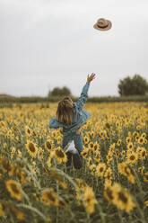 Rückansicht einer jungen Frau mit blauer Jeansjacke, die einen Hut in ein Sonnenblumenfeld wirft - OCAF00432