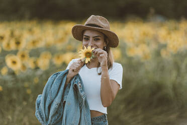 Porträt einer jungen Frau mit blauer Jeansjacke und Hut in einem Sonnenblumenfeld - OCAF00434