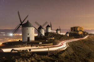 Spain, Province of Toledo, Consuegra, Vehicle light trails in front of row of old windmills standing on top of hill at night - WPEF02112