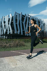 Junge Frau joggt auf einem Weg, modernes Gebäude im Hintergrund - MTBF00041