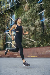 Side view of young female jogger running in a city with a garden facade building in the background - MTBF00046