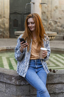 Woman using smartphone sitting on a fountain - AFVF04148