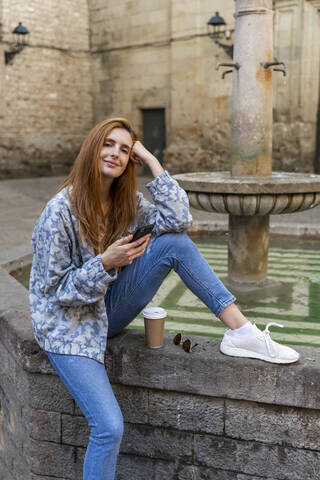 Frau mit Smartphone auf einem Springbrunnen sitzend, lizenzfreies Stockfoto