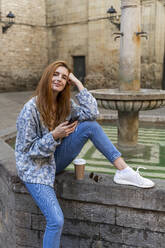 Woman using smartphone sitting on a fountain - AFVF04149