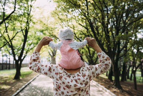Mutter, die mit ihrem kleinen Mädchen auf den Schultern in einem Park spazieren geht - GEMF03235