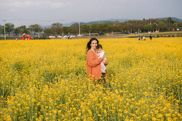Porträt einer glücklichen Frau mit einem kleinen Mädchen auf dem Arm in einem Rapsfeld, Gyeongju, Südkorea - GEMF03238