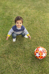 Porträt eines lächelnden kleinen Jungen mit einem Ball auf dem Rasen hockend - VGF00316