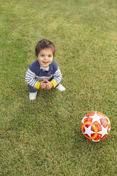 Porträt eines glücklichen kleinen Jungen mit einem Ball auf dem Rasen hockend - VGF00317