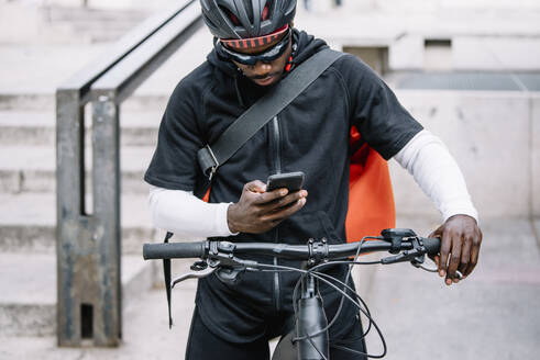 Stylischer junger Mann mit Fahrrad, Smartphone und Umhängetasche in der Stadt - CJMF00156