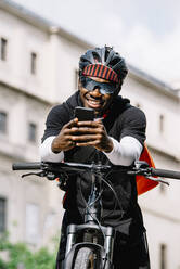 Stylischer junger Mann mit Fahrrad, Smartphone und Umhängetasche in der Stadt - CJMF00158