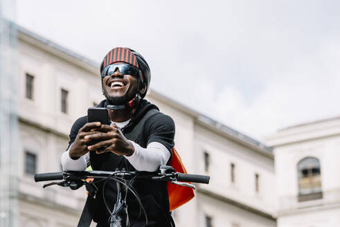 Glücklicher stilvoller junger Mann mit Fahrrad, Smartphone und Umhängetasche in der Stadt - CJMF00159